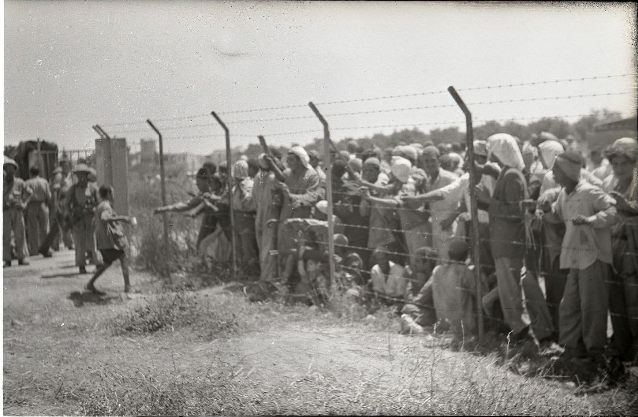 📷 Историческое чёрно-белое фото (Палестина 1948 г.): большая толпа арабских людей — мужчины, женщины, дети — стоят и сидят вдоль ограждения из колючей проволоки; охранник слева.