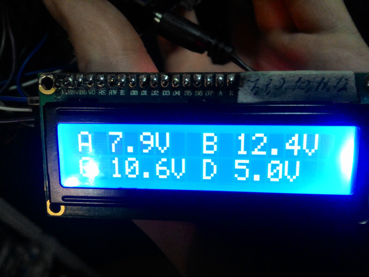 Фото крупным планом синего LCD-дисплея 16×2 в руке: на экране две строки — «A 7.9V  B 12.4V» и «C 10.6V D 5.0V» (показания напряжений четырёх каналов)
