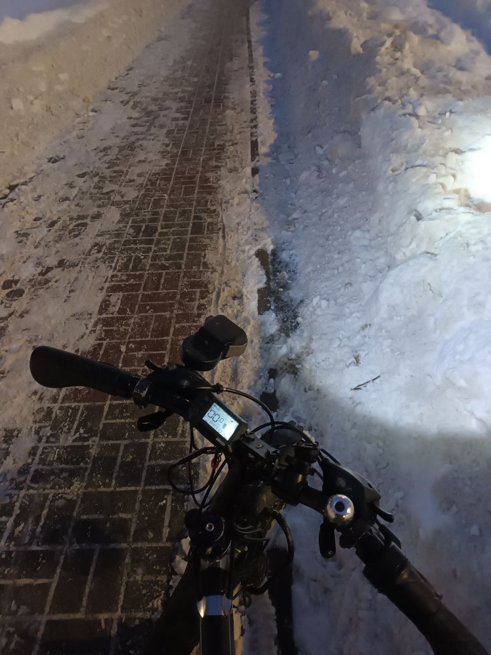 📷 Вид с велосипеда ночью: руль с дисплеем, заснеженная тротуарная плитка, снежный бордюр.