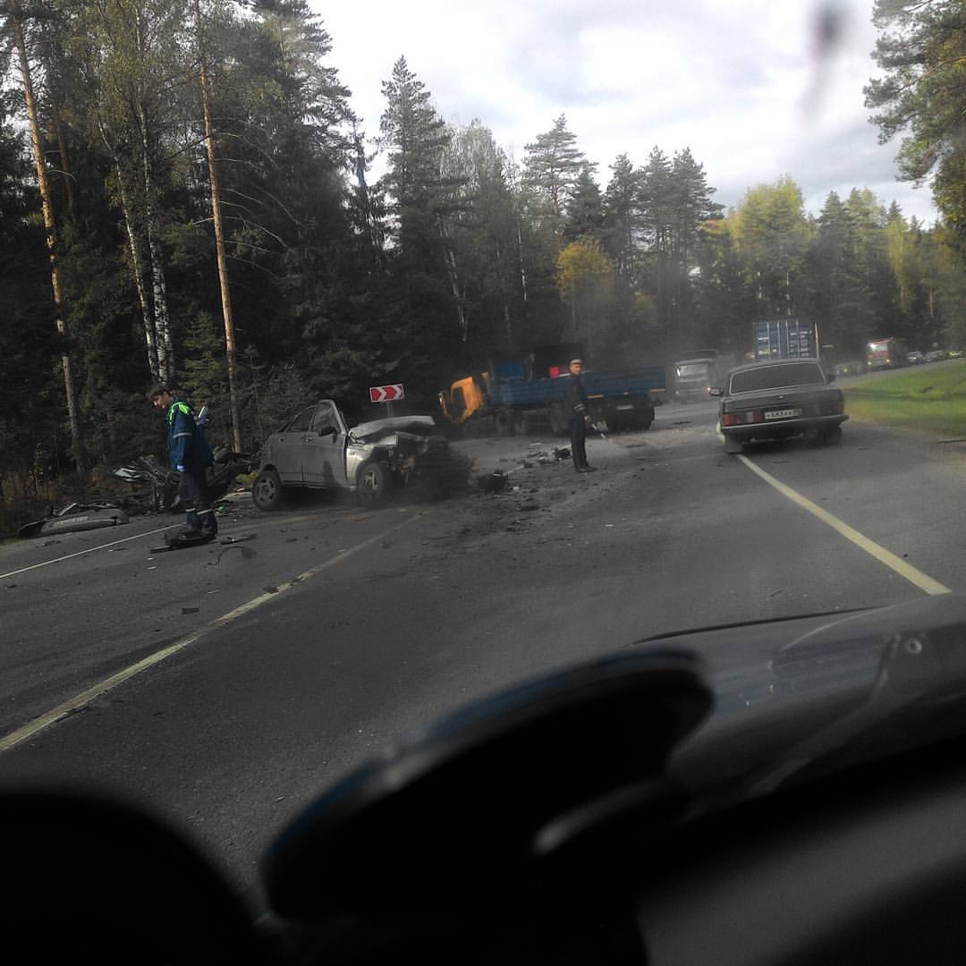 Вид из салона автомобиля через лобовое стекло на место серьёзного ДТП: на дороге — сильно разбитый белый автомобиль, обломки и мусор на асфальте, синий грузовик с поднятым кузовом; вокруг стоят люди (предположительно аварийные службы и прохожие); справа объезжает серый седан; по обочинам — берёзы и ели, небо пасмурное, облачное