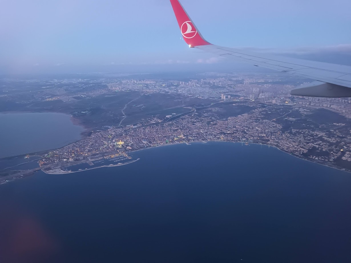 📷 Вид из иллюминатора самолёта Turkish Airlines: крыло с красным логотипом; внизу — береговая линия с городом на морском заливе, полуостров с исторической застройкой; на горизонте — многоэтажная застройка; вечернее синеватое освещение.