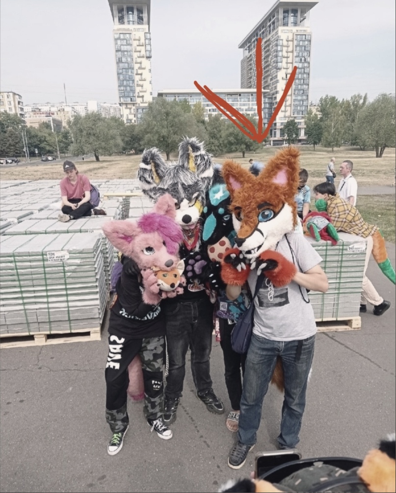 📷 Трое людей с фурсьютами (розовый, серый с бирюзовым, рыжий лис) на улице у многоэтажек, красная стрелка указывает на среднего в рыжем фурсьюте.