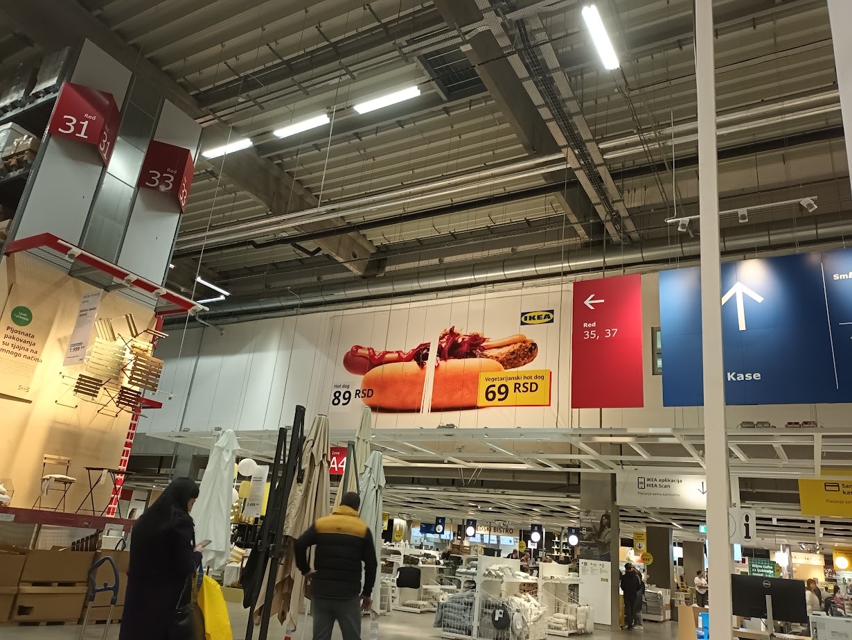 📷 Внутри торгового центра IKEA: ряды с подвешенными ценниками; красный подвешенный объект (упаковка-подарок); синяя стрела-указатель «Kasse» (касса); высокий металлический потолок.