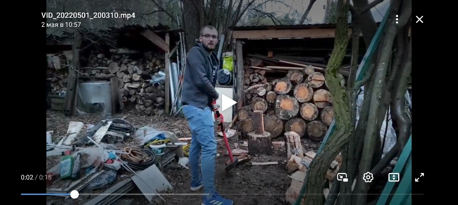 📋 Скриншот видео (VID_20220501_200310.mp4, 2 мая в 10:57). Мужчина стоит во дворе/сарае среди дров, мусора и инструментов. Продолжительность 0:18.