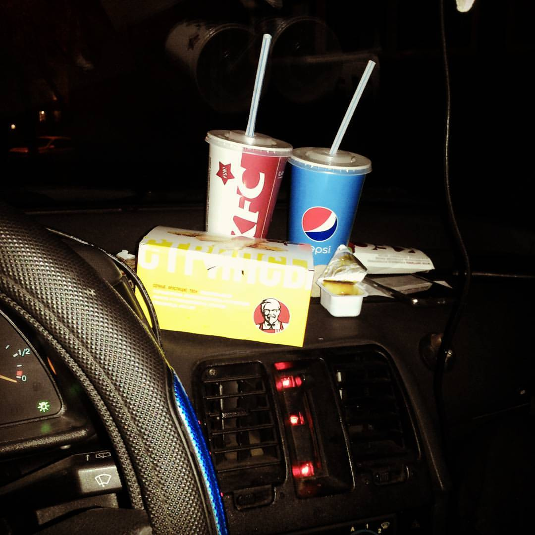 Ночной интерьер машины: на приборной панели стоят красный стакан KFC с логотипом и синий стакан Pepsi с трубочками; перед ними — жёлтая коробка KFC с логотипом полковника Сандерса; слева — рулевое колесо; подсветка приборной панели красная
