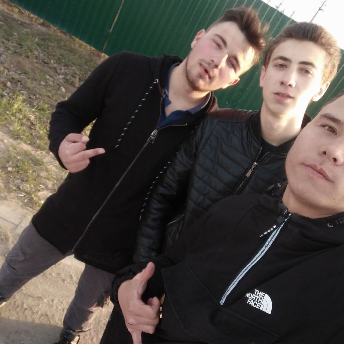 📷 Трое молодых парней делают сэлфи на улице у зелёного забора — двое в чёрных куртках, один на переднем плане в куртке The North Face.