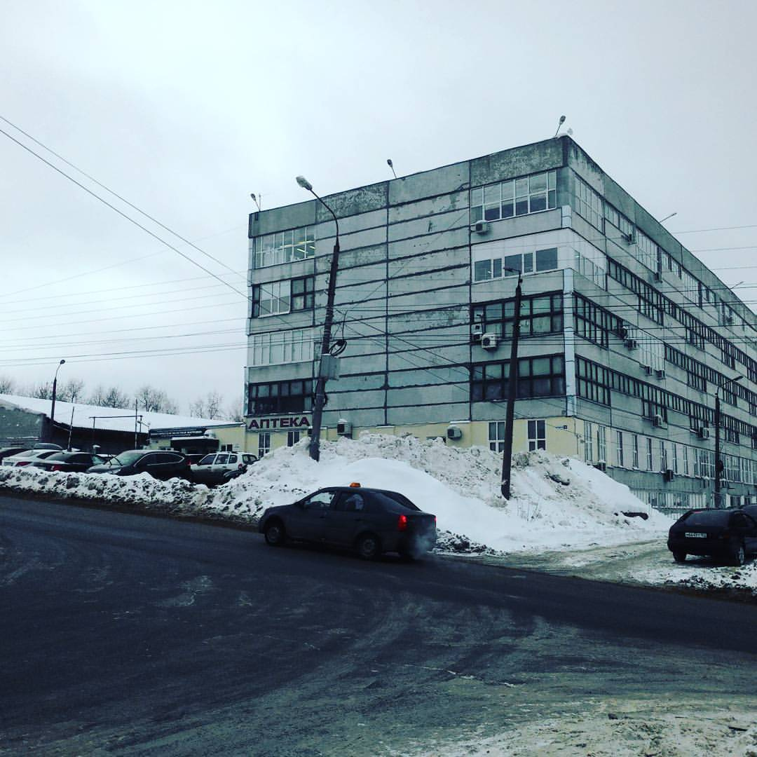 Фото зимней улицы: советское 5-этажное здание с фасадом из стекла и бетона, у входа вывеска «АПТЕКА»; снег, припаркованные машины (чёрный седан на переднем плане); пасмурное серое небо; дорога на переднем плане