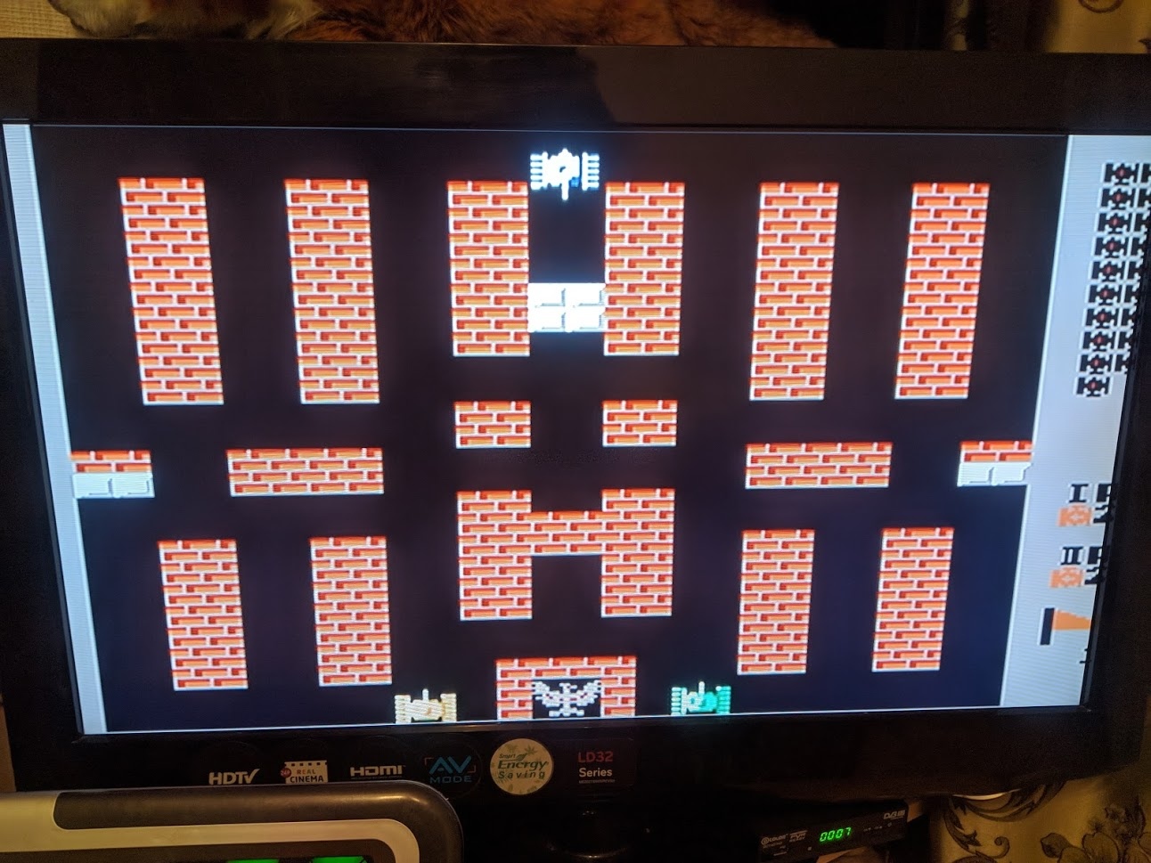 Телевизор с надписью HDV на корпусе: на экране игра Battle City («Танчики», Dendy/Famicom) — классический уровень с лабиринтом из красных кирпичных стен, белый квадратик-танк; справа боковая информационная панель игры