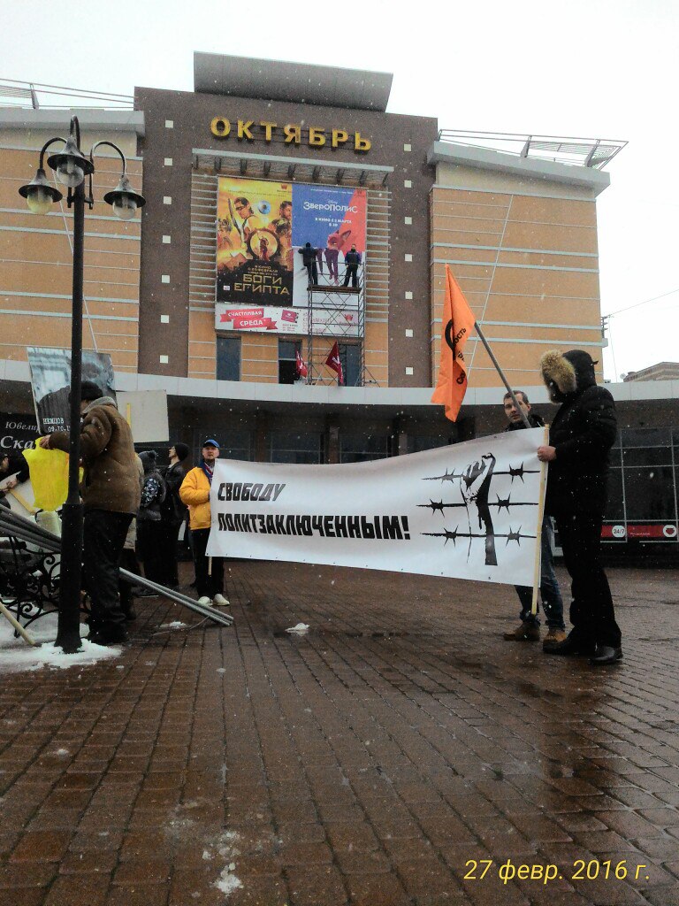 Фото в снегопад у кинотеатра «Октябрь»: несколько активистов держат горизонтальный баннер «СВОБОДУ ПОЛИТЗАКЛЮЧЁННЫМ!» с изображением человека за колючей проволокой, рядом оранжевый флаг; на фасаде кинотеатра афиши — «Зверополис» и другие фильмы; дата на фото «27 февр. 2016 г.»