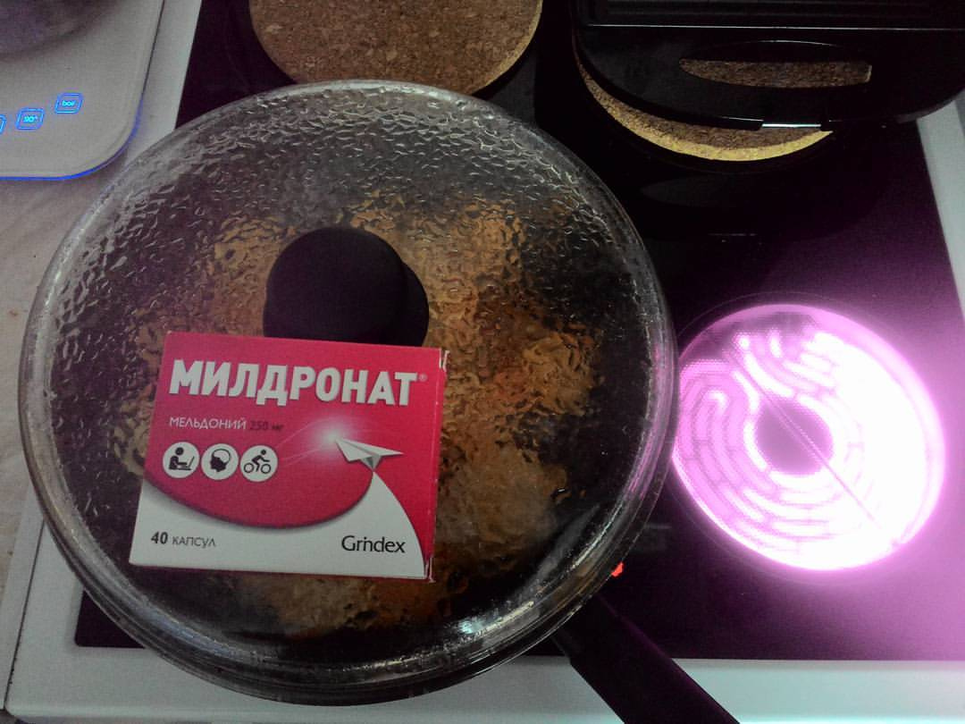 Стеклянная крышка сковороды на электроплите; на крышке лежит красная картонная коробка с надписью «МИЛДРОНАТ / Мельдоний / 40 капсул / Grindex» и пиктограммами спортсменов; справа на плите светится раскалённая конфорка