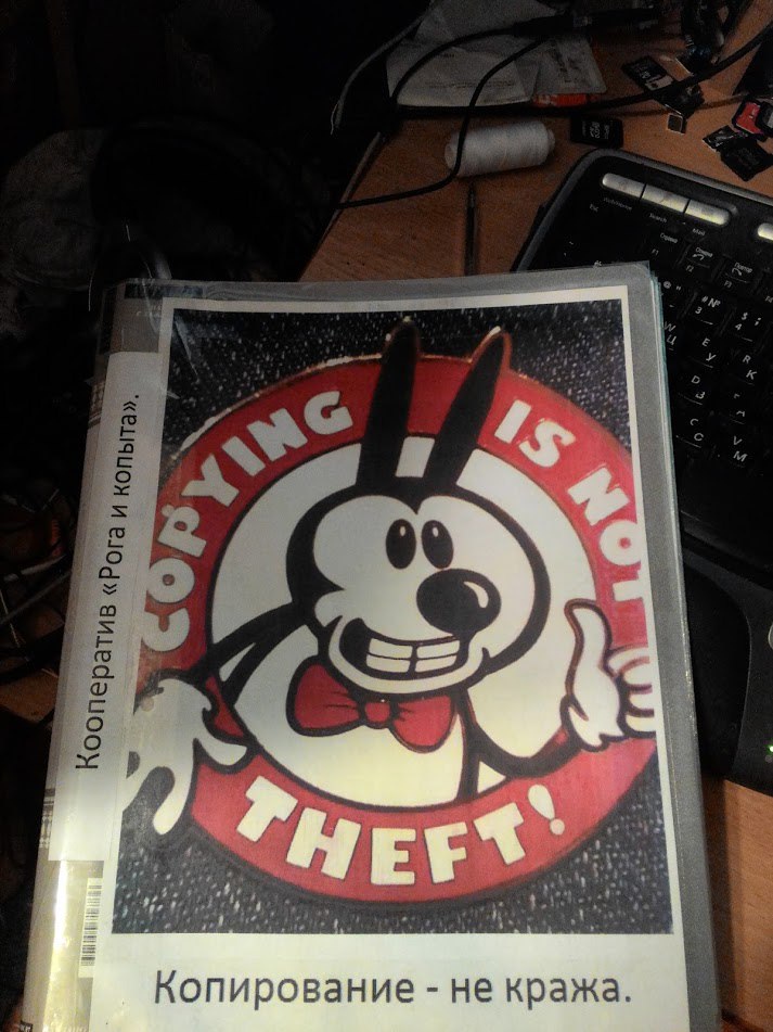 Стикер в пластиковой папке: круглый логотип «COPYING IS NOT THEFT!» с мультяшным персонажем в галстуке-бабочке; подпись на корешке папки «Кооператив «Рога и копыта»»; под стикером надпись «Копирование — не кража.»; позади клавиатура и провода