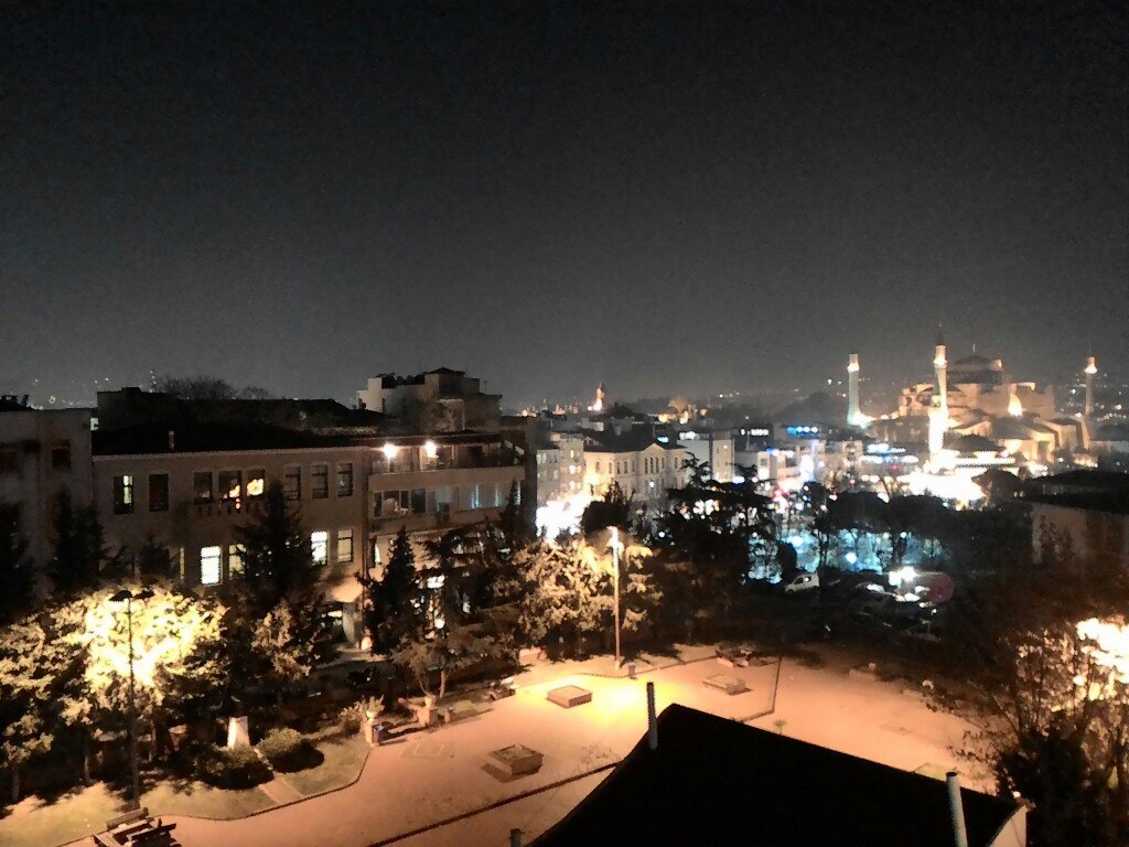 Ночная панорама Стамбула из окна гостиницы: на переднем плане — заснеженный парк с деревьями и фонарями; вдали освещены купол и минарет Айя-Софии (справа); тёмное небо, городские огни