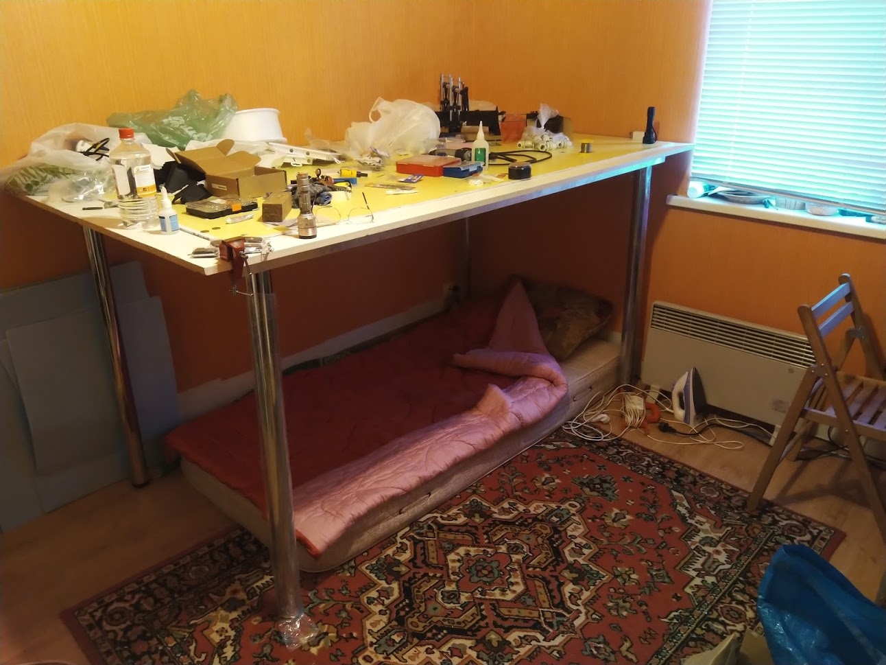 📷 Комната с оранжевыми стенами: большой белый рабочий стол/верстак, захламлённый инструментами и электроникой. Под столом прямо на полу — матрас с подушкой. Ковёр, окно.