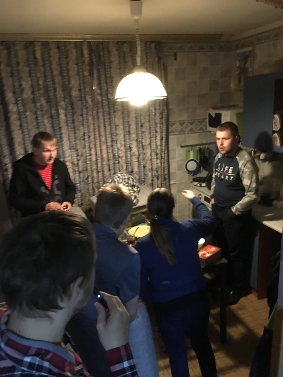 📷 Несколько молодых людей в небольшой квартире вечером: двое слева сидят, один справа в футболке «LIFE IS ART» стоит жестикулируя, несколько других стоят рядом, над ними плафон, кухонный интерьер.