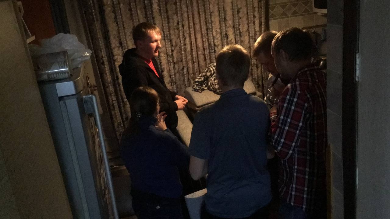 📷 Группа людей в тёмном коридоре — несколько человек склонились над диваном, один в красно-чёрной куртке стоит слева.