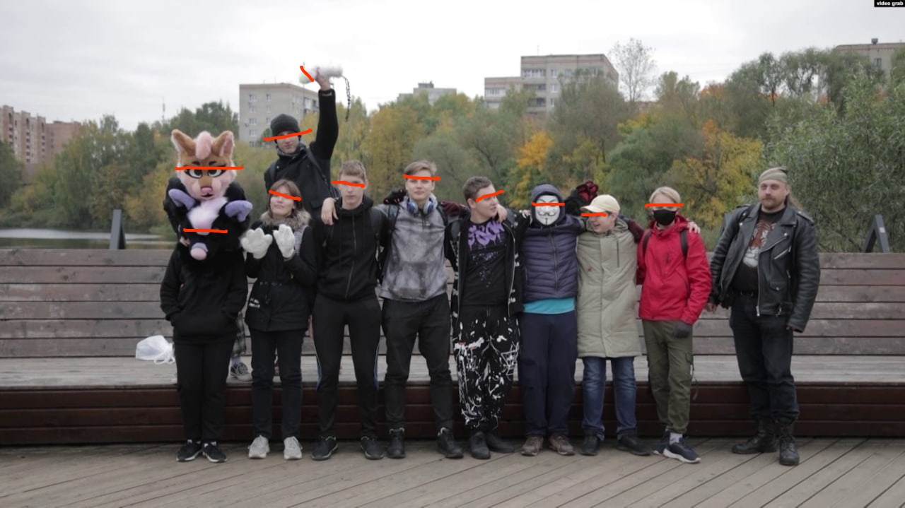 📷 Группа из ~10 человек на деревянном настиле у реки на фоне осенних деревьев и многоэтажных домов — слева человек в голове поросёнка (фурсьют-маска), рядом дети/подростки и взрослые, обнимаются, лица отмечены оранжевыми полосками. Скриншот из видео (video.grab).
