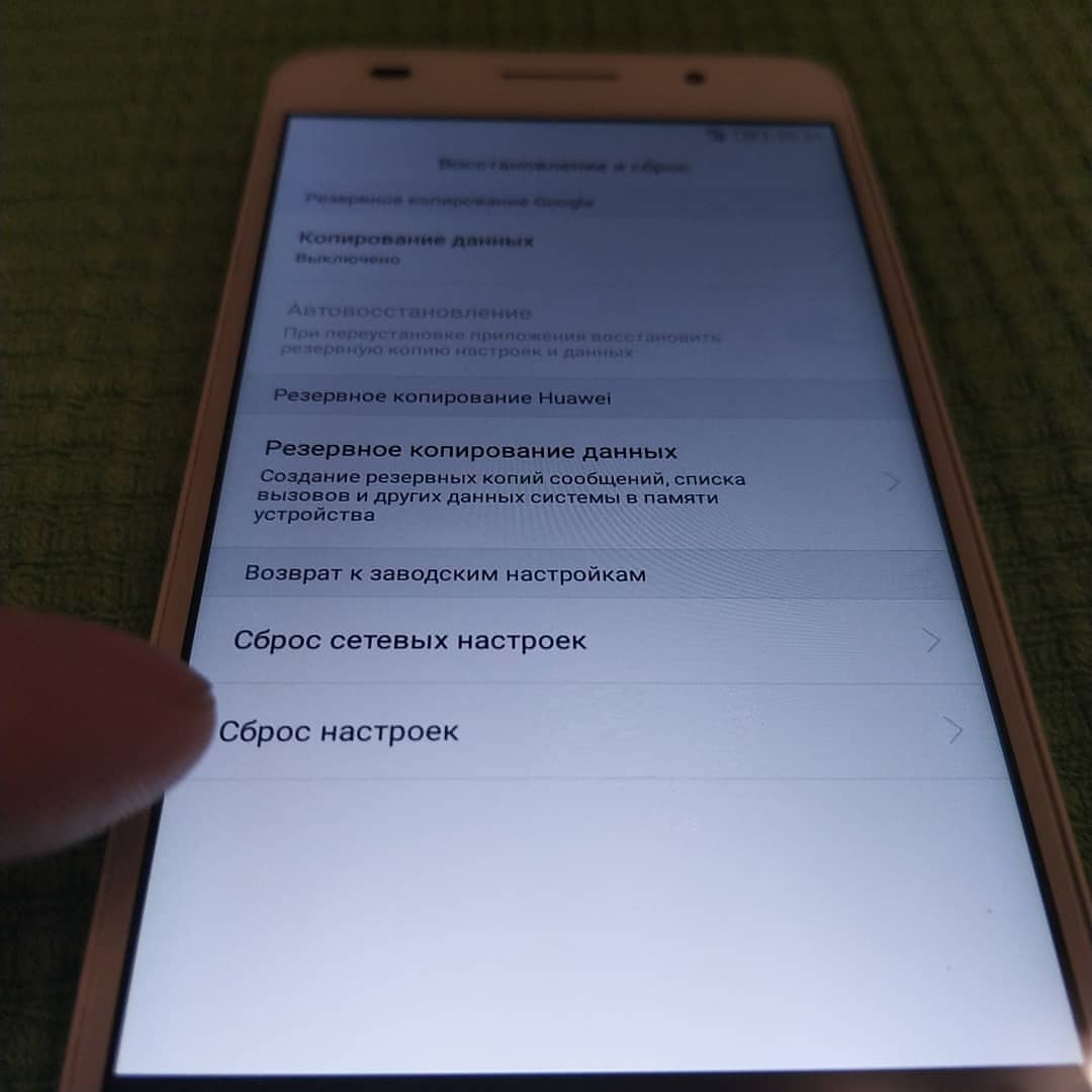 Смартфон Huawei Honor 6 лежит на зелёной поверхности; экран открыт на разделе «Восстановление и сброс»: Резервное копирование Google / Копирование данных — Выключено / Автовосстановление / Резервное копирование Huawei / Резервное копирование данных / Возврат к заводским настройкам / Сброс сетевых настроек / Сброс настроек; палец касается пункта «Сброс настроек»