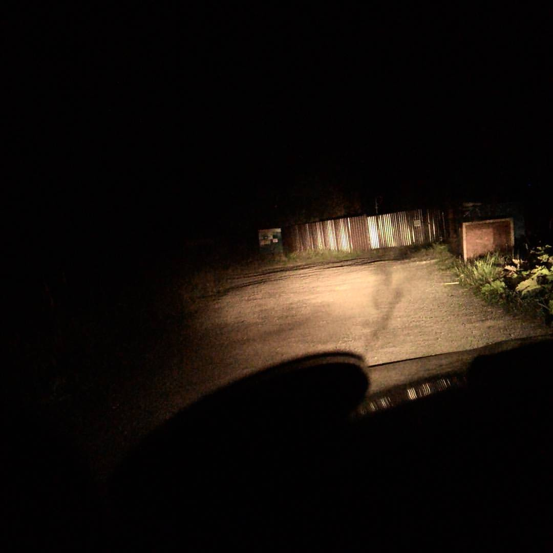 Ночная поездка: вид из тёмного салона автомобиля — в свете фар едва видна грунтовая дорога; справа — гофрированный металлический забор, подсвеченный фарами, и тёмное кирпичное строение; вокруг полная темнота