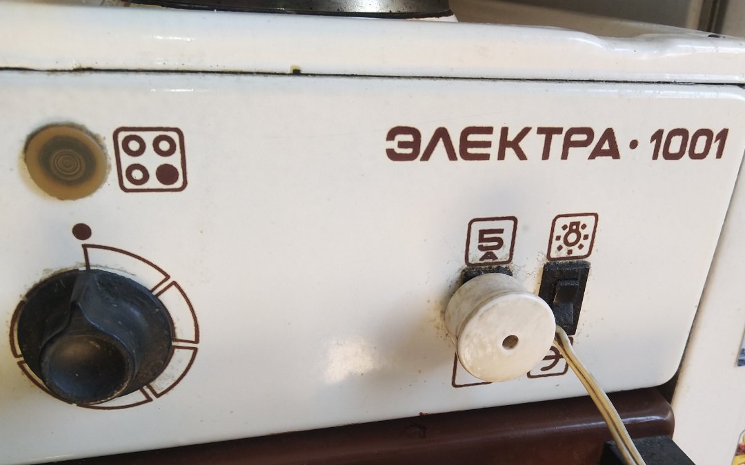 Крупный план передней панели советской электрической плиты «Электра 1001» белого цвета с коричневым декором: слева — крупная чёрная поворотная ручка регулятора с пиктограммой конфорок; справа — надпись «ЭЛЕКТРА · 1001» и два тумблера с белыми фарфоровыми рукоятками; над тумблерами — пиктограмма гриля/вертела и значок подсветки