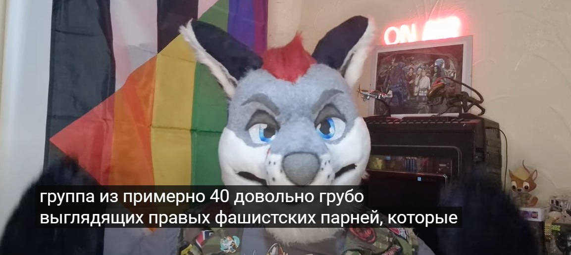 📋 Скриншот видео: фурсьют серо-голубого волка/зайца перед радужным флагом и флагом прогресс-прайда; на экране «ON AIR»; субтитры внизу: «группа из примерно 40 довольно грубо выглядящих правых фашистских парней, которые»