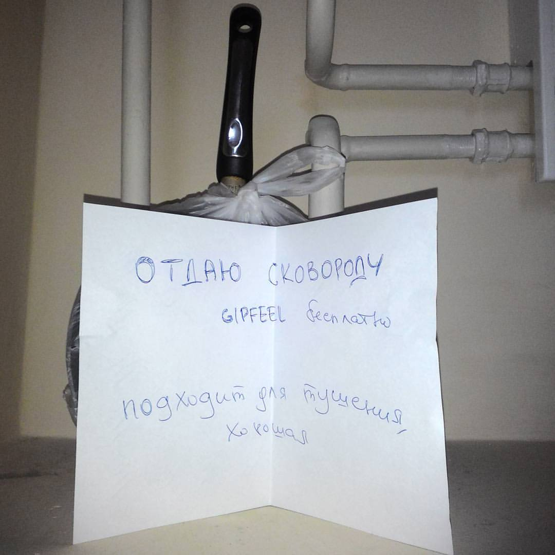 Фото в подъезде у серых труб: рукописная записка на белом листе бумаги: «ОТДАЮ СКОВОРОДУ / GIPFEEL бесплатно / подходит для тушения / хо хорошая!!»; за запиской прислонена сковорода с чёрной ручкой, завёрнутая в пакет