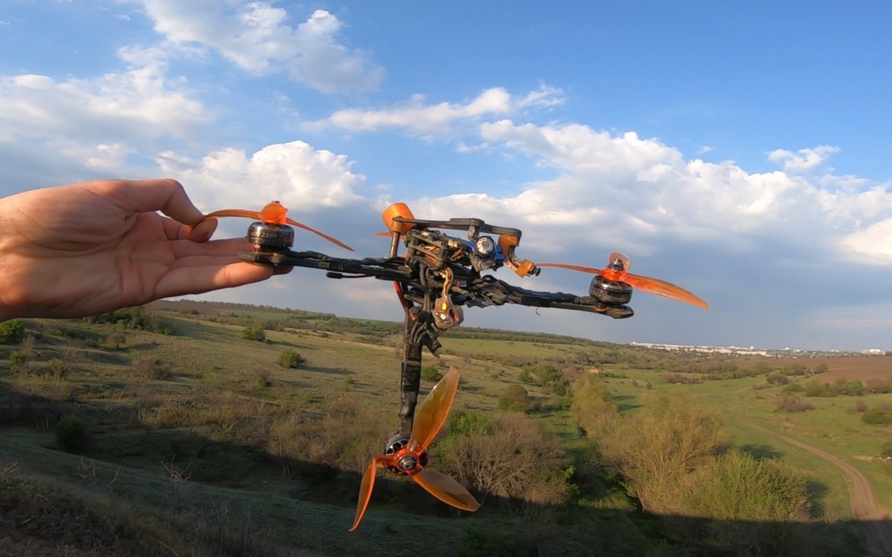Гоночный FPV-квадрокоптер с оранжевой рамой и оранжевыми пропеллерами на руке; фон — зелёный луг, синее небо с облаками