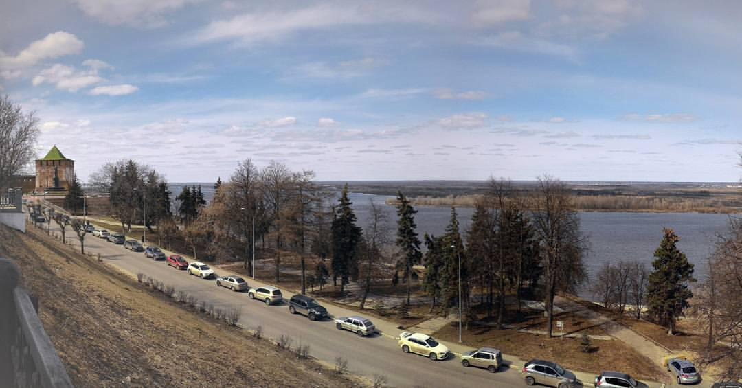 Панорамное фото Нижнего Новгорода: слева башня Нижегородского кремля с зелёной кровлей и белыми стенами, ниже — дорога с припаркованными автомобилями вдоль берега, деревья без листьев, справа — широкая Волга, вдали противоположный берег; небо с облаками, ранняя весна