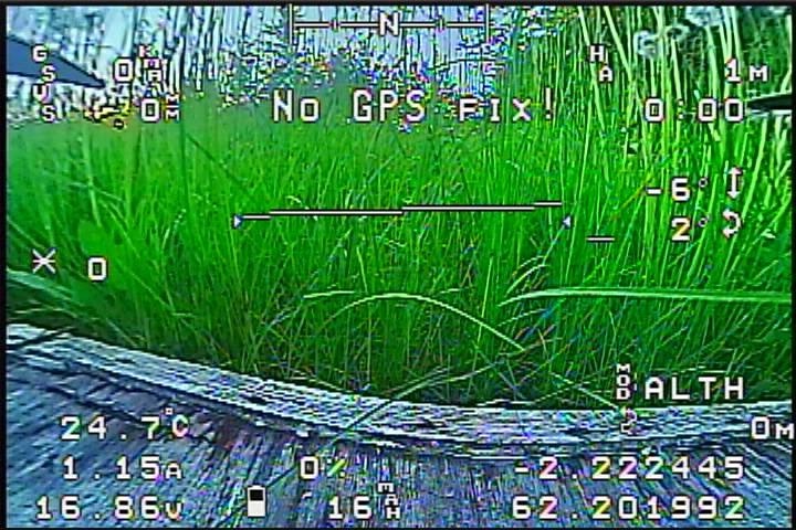 Скриншот FPV-камеры дрона: зелёная трава крупно, по центру красная надпись «No GPS Fix!»; OSD-данные: «24 7°C / 1.15A / 16.86V / 0% / 16° / -2.222445 / 62.201992 / ALTH / -0M»; N на компасе