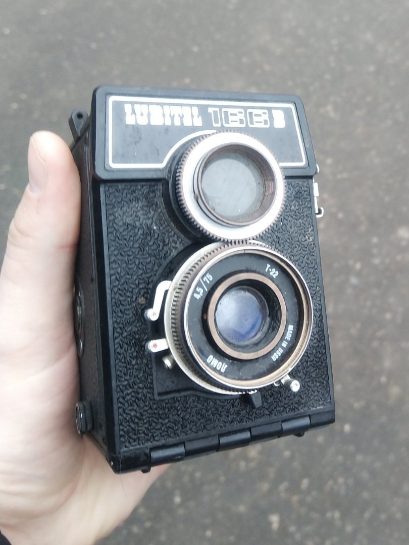 Советский двухобъективный зеркальный фотоаппарат «LUBITEL 166Б» (Любитель 166Б) в руке на фоне мокрого асфальта; два объектива — видоискательный и съёмочный, надпись «made in USSR» на чёрном корпусе