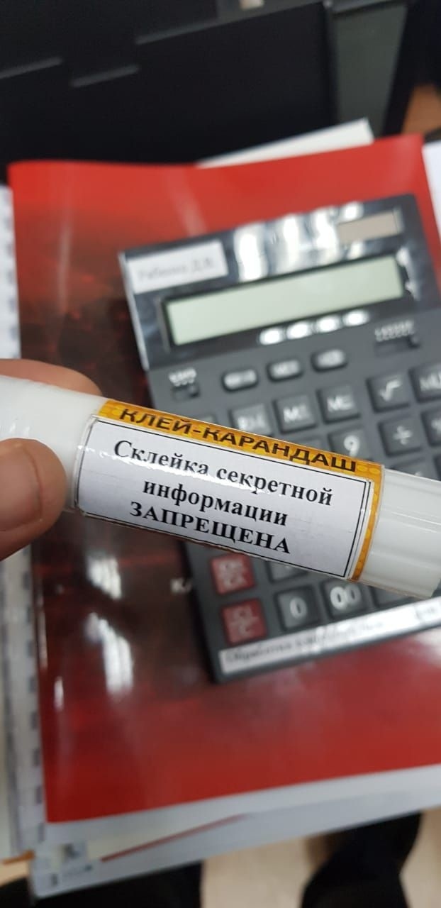 Клей-карандаш «КЛЕЙ-КАРАНДАШ» с наклейкой «Склейка секретной информации ЗАПРЕЩЕНА»; на заднем плане калькулятор и красные папки