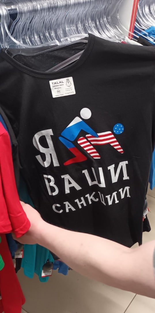 🖼️ Чёрная футболка в магазине: принт — два стикмена в цветах российского и американского флагов в пошлой позе; текст «Я [фигурка] ВАШИ САНКЦИИ»; бирка «TALAL».