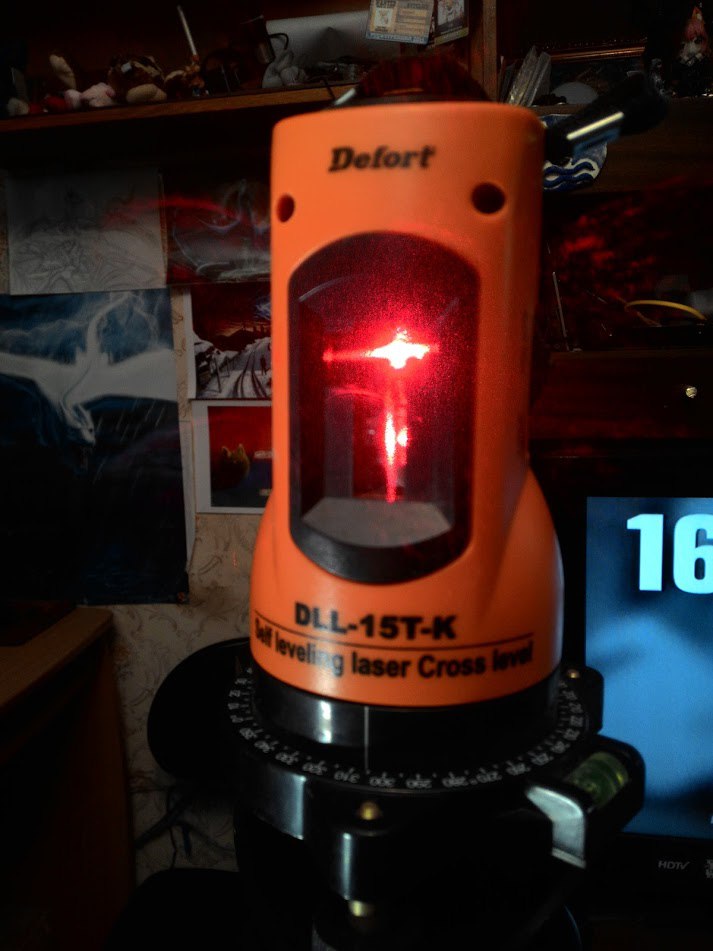 Оранжевый лазерный нивелир Defort DLL-15T-K (Self Leveling Laser Cross Level) крупным планом: в окошке горит красный лазерный крест; на заднем плане монитор с цифрой «16», полки с вещами и плакаты