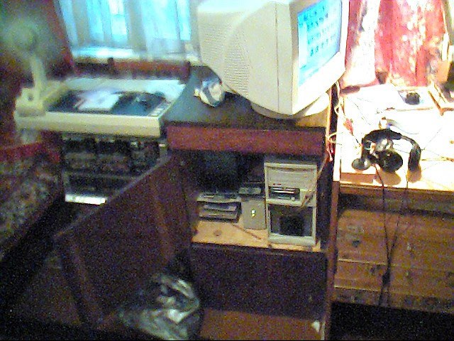 Старый компьютерный уголок ~2004 года: CRT-монитор (синий экран), системный блок, VHS-плеер или кассеты; справа дисковый телефон на деревянной полке; диван с ковровой обивкой, окно с занавеской на заднем плане