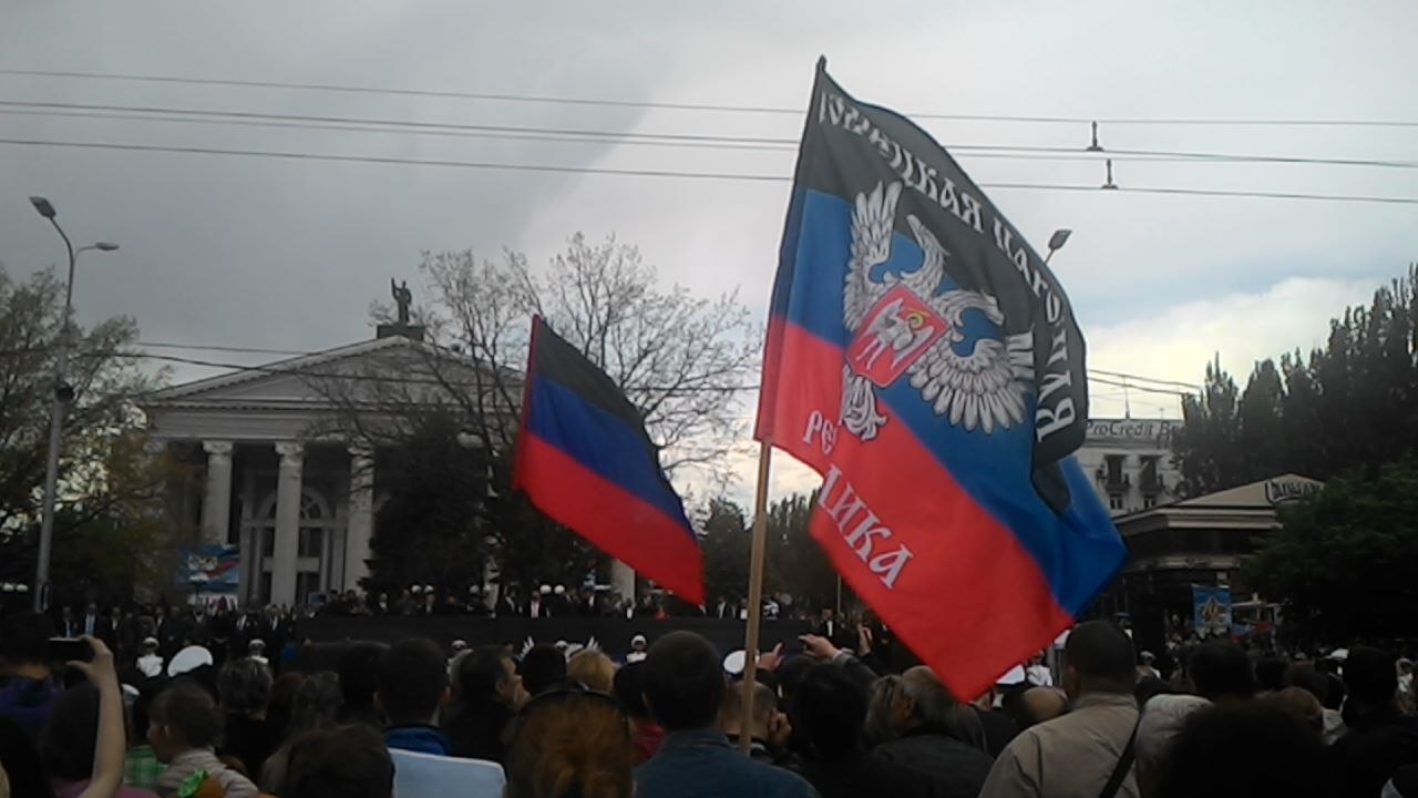 Митинг в Донецке: толпа людей, над которой развеваются флаги ДНР и чёрное полотнище с надписью и орлом; на заднем плане колонный портик театра и деревья