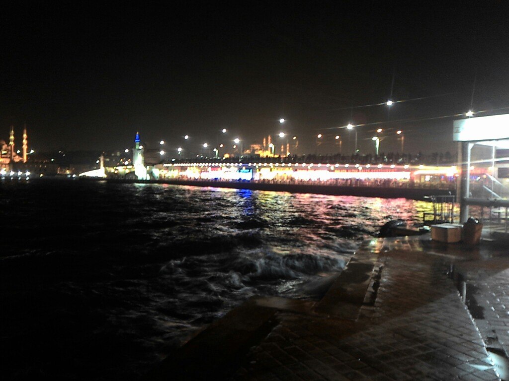 Вид на Галатский мост и Золотой Рог ночью: тёмная бурлящая вода залива, мост освещён рядами огней, отражения в воде; вдали слева — мечеть, справа — причал с навесом