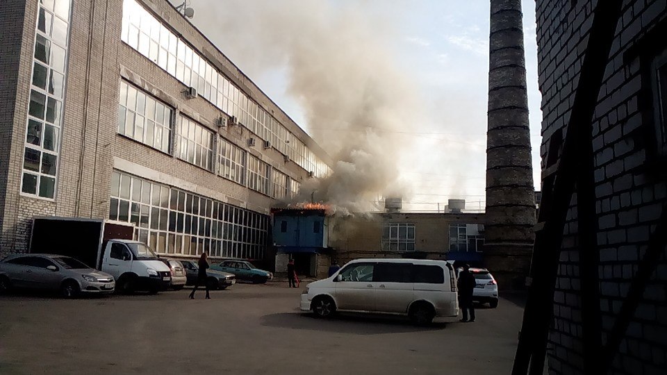 Пожар: крупное промышленное здание, дым и огонь вырываются с крыши одноэтажной пристройки; несколько человек стоят во дворе, припаркованы автомобили