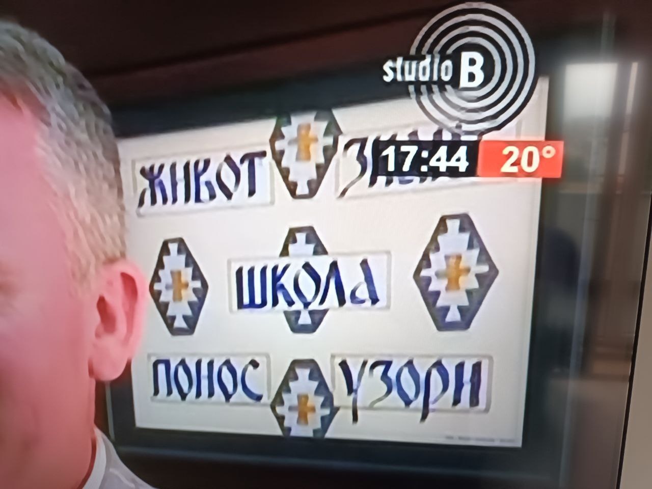 📷 Экран телевизора канала Studio B (17:44, 20°): надписи в стилизованном шрифте с крестообразными символами — «ЖИВОТ», «ШКОЛА», «ПОНОС», «УЗОРИ»; рядом видна часть головы мужчины.