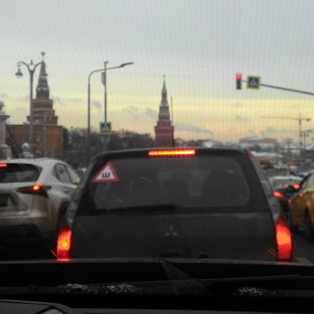 Вид из салона автомобиля на пробку у Кремля в Москве: прямо впереди — тёмный Mitsubishi с наклейкой «Ш» (шипованные шины); на горизонте между машинами — красно-кирпичные башни Московского Кремля; справа — красный светофор и дорожный знак «пешеходный переход»; правее — жёлтое такси; вечернее небо с розово-оранжевым закатом