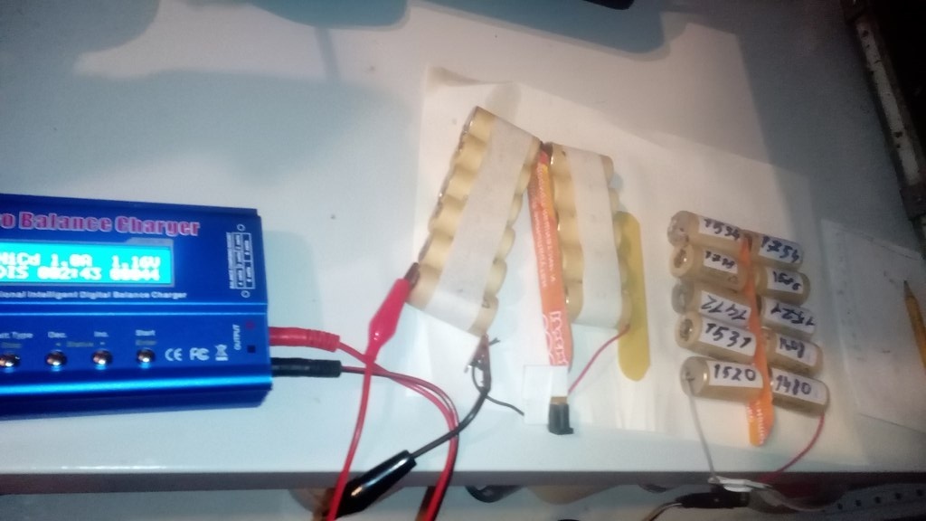 Balance Charger с красными зажимами заряжает самодельный аккумуляторный блок; рядом 12 цилиндрических элементов с наклейками, подписанными маркером: 1536, 1355, 1738, 1108, 1531, 1100, 1520, 1470