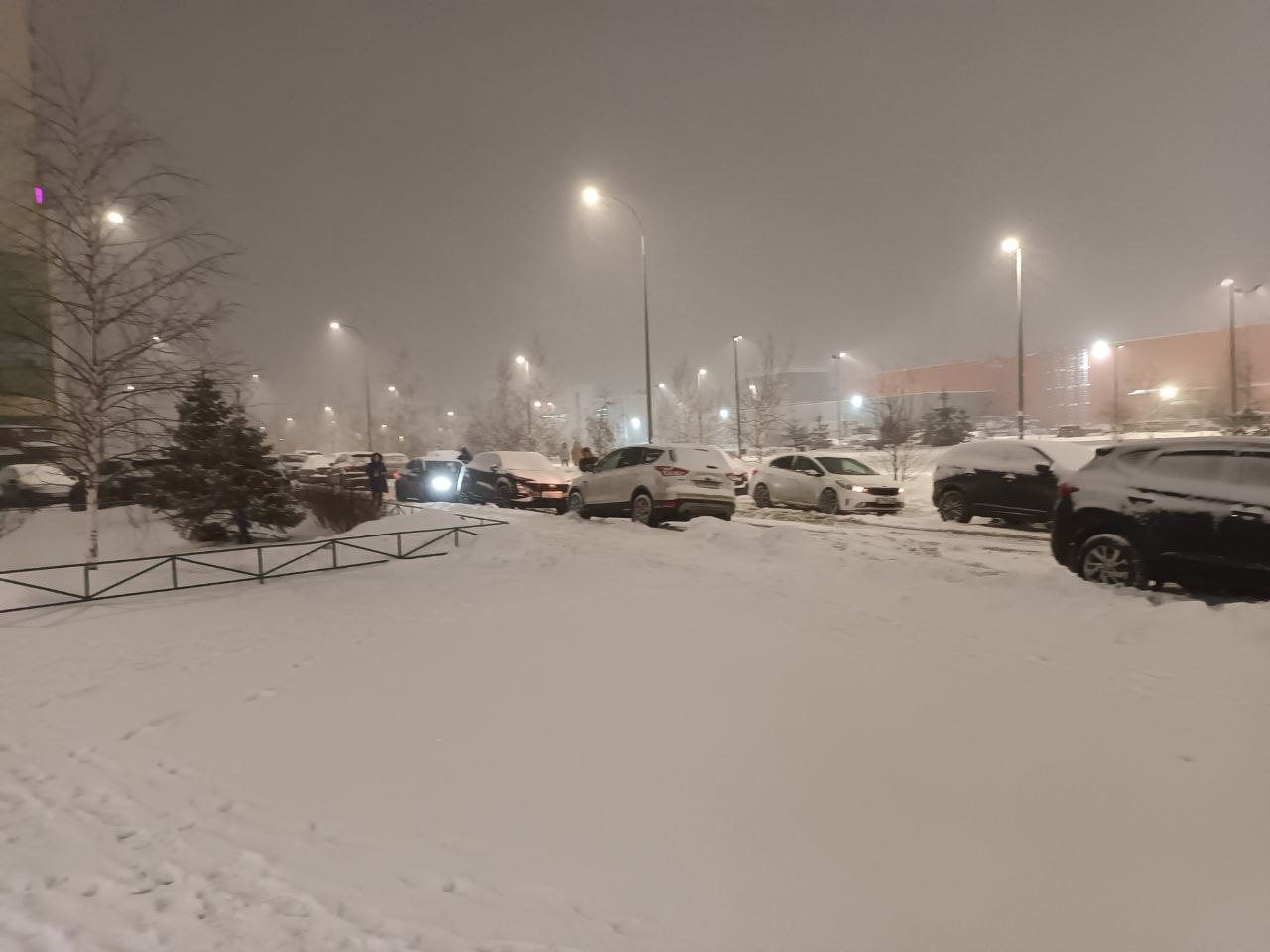 📷 Ночная снежная парковка: несколько автомобилей, фонари, туман, вдали здание торгового центра.