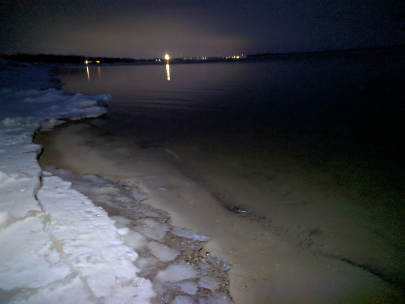 Ночной берег реки/водоёма зимой: лёд и снег у кромки берега, тёмная вода; огни и отражения на другом берегу, тёмное небо