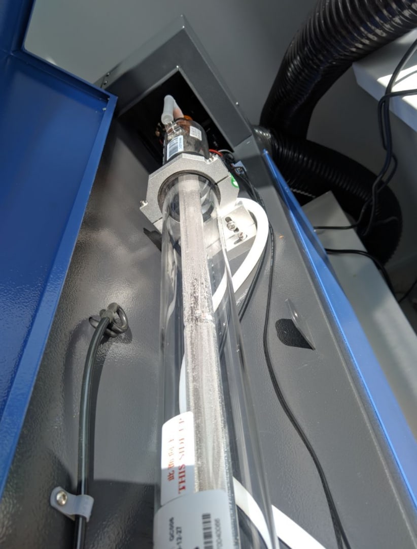 📷 Стеклянная CO₂-лазерная трубка внутри синего металлического корпуса станка: белые трубки системы охлаждения, гофрированный вентиляционный шланг вверху, металлический держатель