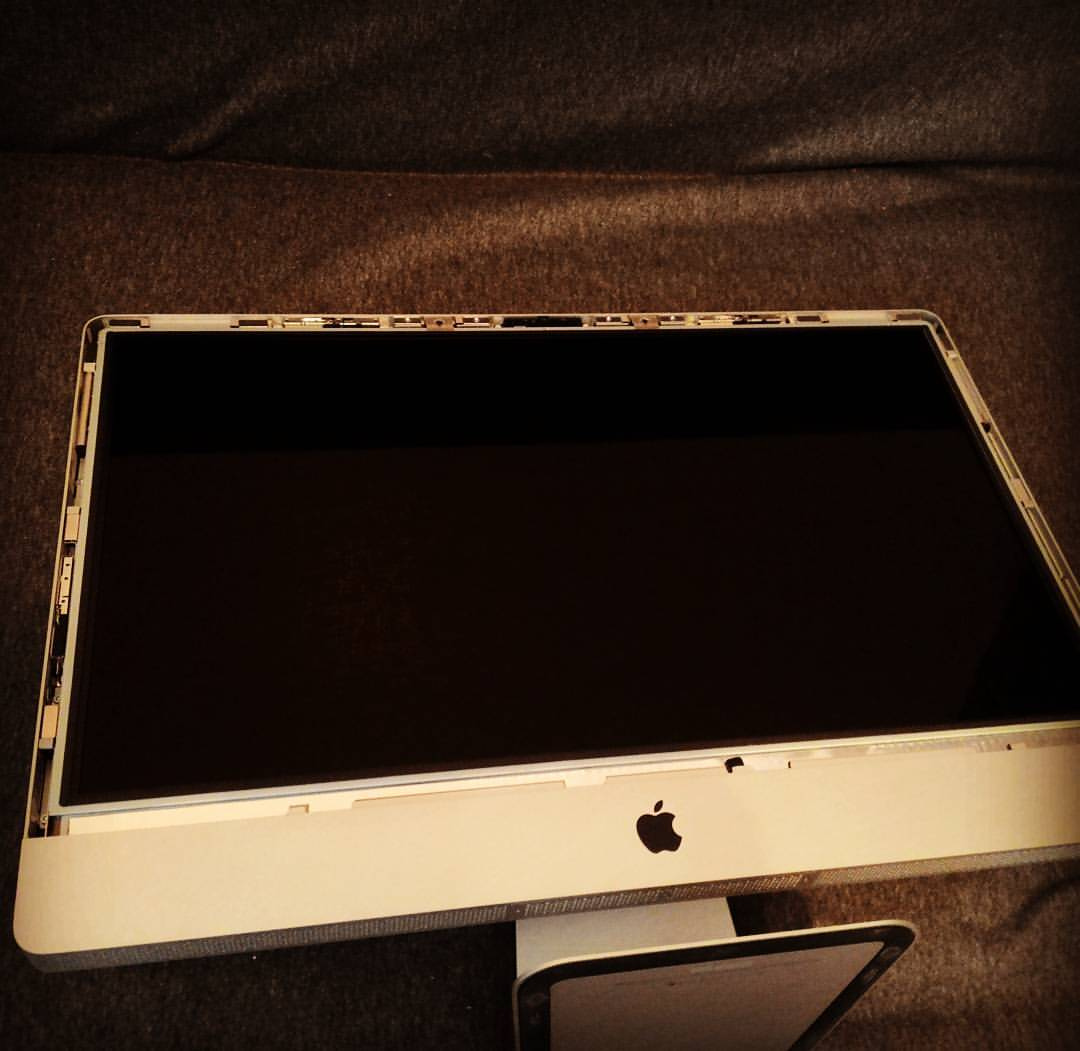 Экран Apple iMac (LCD-матрица с алюминиевой и пластиковой рамкой-безелем) снят с корпуса и лежит лицевой стороной вверх на тёмной тканевой поверхности (диван или кровать); экран чёрный, выключен; в нижней части рамки виден логотип Apple; по верхнему краю — внутренний каркас с разъёмами