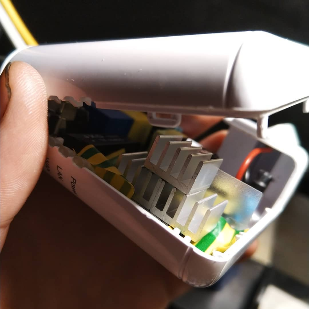 Рука держит раскрытый белый роутер (MikroTik): хорошо видны внутренности — серебристый радиатор процессора с рёбрами охлаждения по центру, рядом платы, жёлто-зелёные провода, синяя и оранжевая детали; на корпусе надписи «LAN» и «Power»; снято на ярком дневном свету