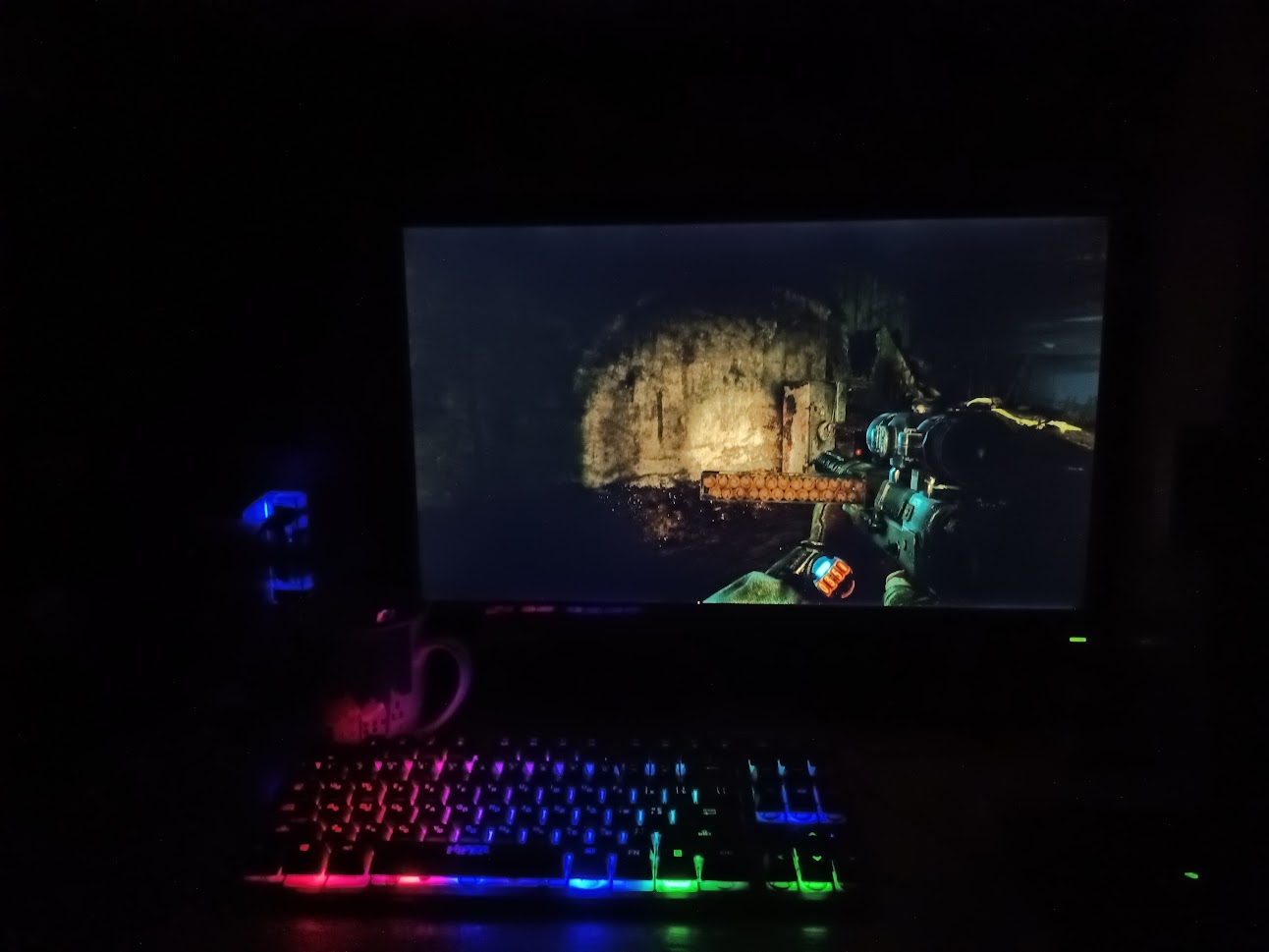 📷 RGB-подсветка клавиатуры в тёмной комнате. На экране монитора — видеоигра FPS (тёмные подземные локации, вид от первого лица, персонаж с оружием). Рядом кружка и мышь.