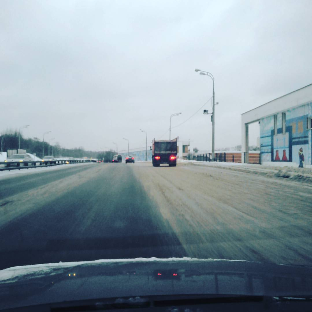 Вид из салона автомобиля через лобовое стекло на зимнюю трассу: мокрый асфальт с остатками снега; прямо впереди — серый грузовик с включёнными красными стоп-сигналами; слева — ещё один автомобиль; справа — современное коммерческое здание; пасмурное серое зимнее небо