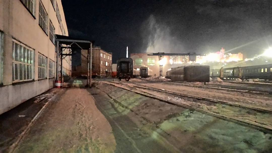 Ночная сцена в вагонном депо: рельсовые пути, покрытые снегом, уходят вдаль; на путях — несколько старых пассажирских вагонов зелёного цвета; слева — большое кирпичное здание депо с освещёнными окнами; вдали — огни и поднимающийся пар; тёмное ночное небо
