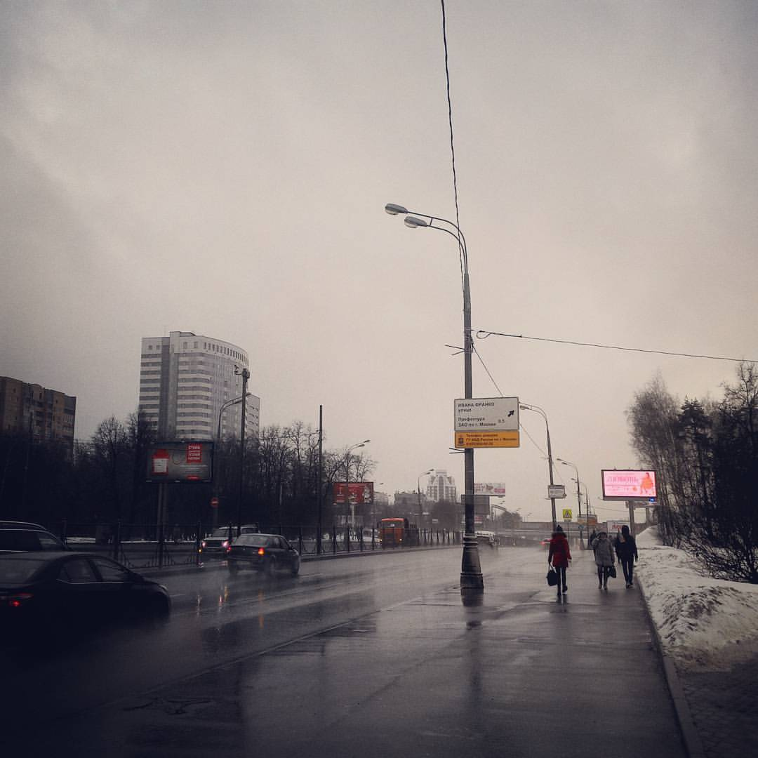 Серая пасмурная московская улица: мокрый широкий проспект с автомобилями, несколько прохожих на тротуаре справа, фонарный столб с жёлтыми дорожными указателями, вдали многоэтажные здания и рекламные щиты, небо затянуто облаками, у дороги остатки снега