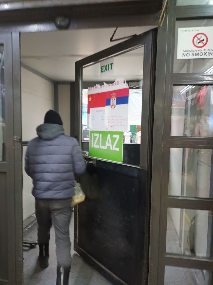 📷 Вход в торговое помещение: дверь с вывеской «EXIT» и зелёной табличкой «IZLAZ»; плакат с флагами Китая и Сербии и надписью на китайском языке; знак «ZABRANJENO PUŠENJE / NO SMOKING»; мужчина в синей куртке выходит.