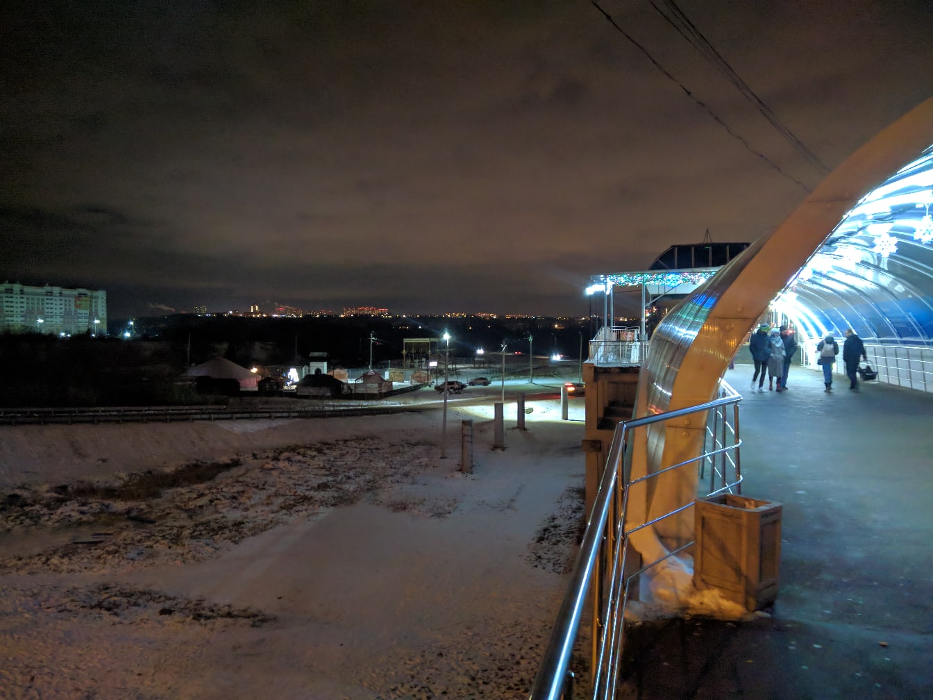 Ночью: современная арка/крытый переход с синей и голубой LED-подсветкой, пешеходы, снег на земле, городские огни и многоэтажки вдали